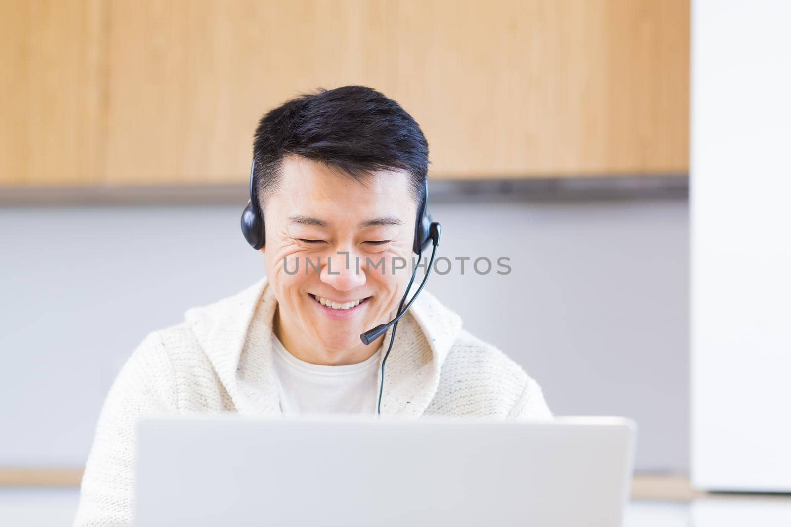 young asian man freelancer sitting at home at office desk with headset and talking online on video link via laptop in room or kitchen. The male indoors in casual clothes does the work and talks