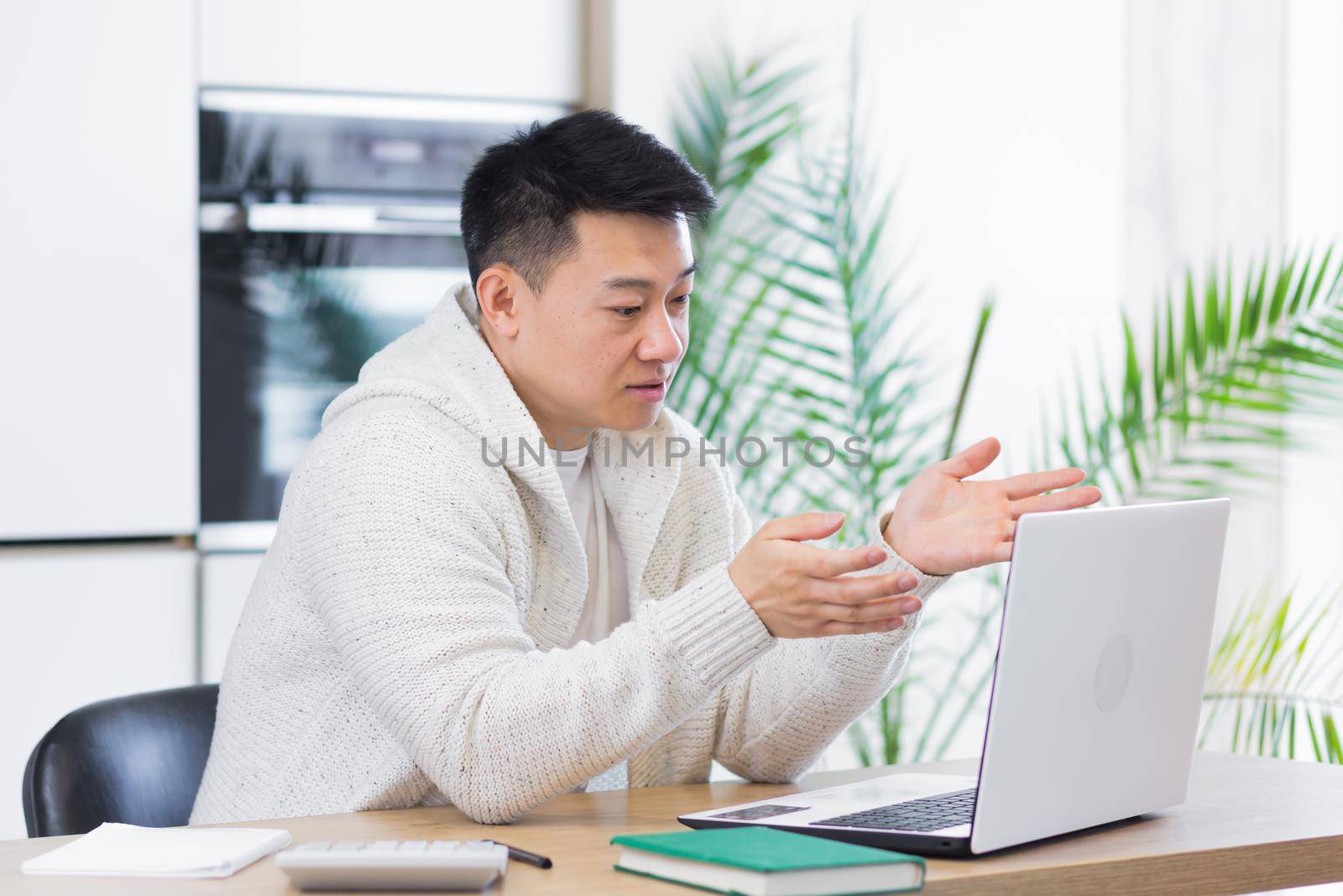 young asian man sitting at home in the living room or kitchen and communicating online. Male waving hand at laptop webcam. Video call learning distance. Training, conference or meeting E-learning