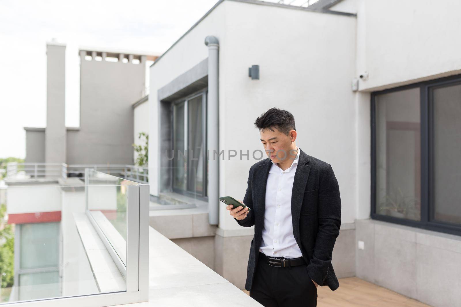 Freelancer Asian businessman in business suit enjoys phone standing on balcony terrace of modern office center
