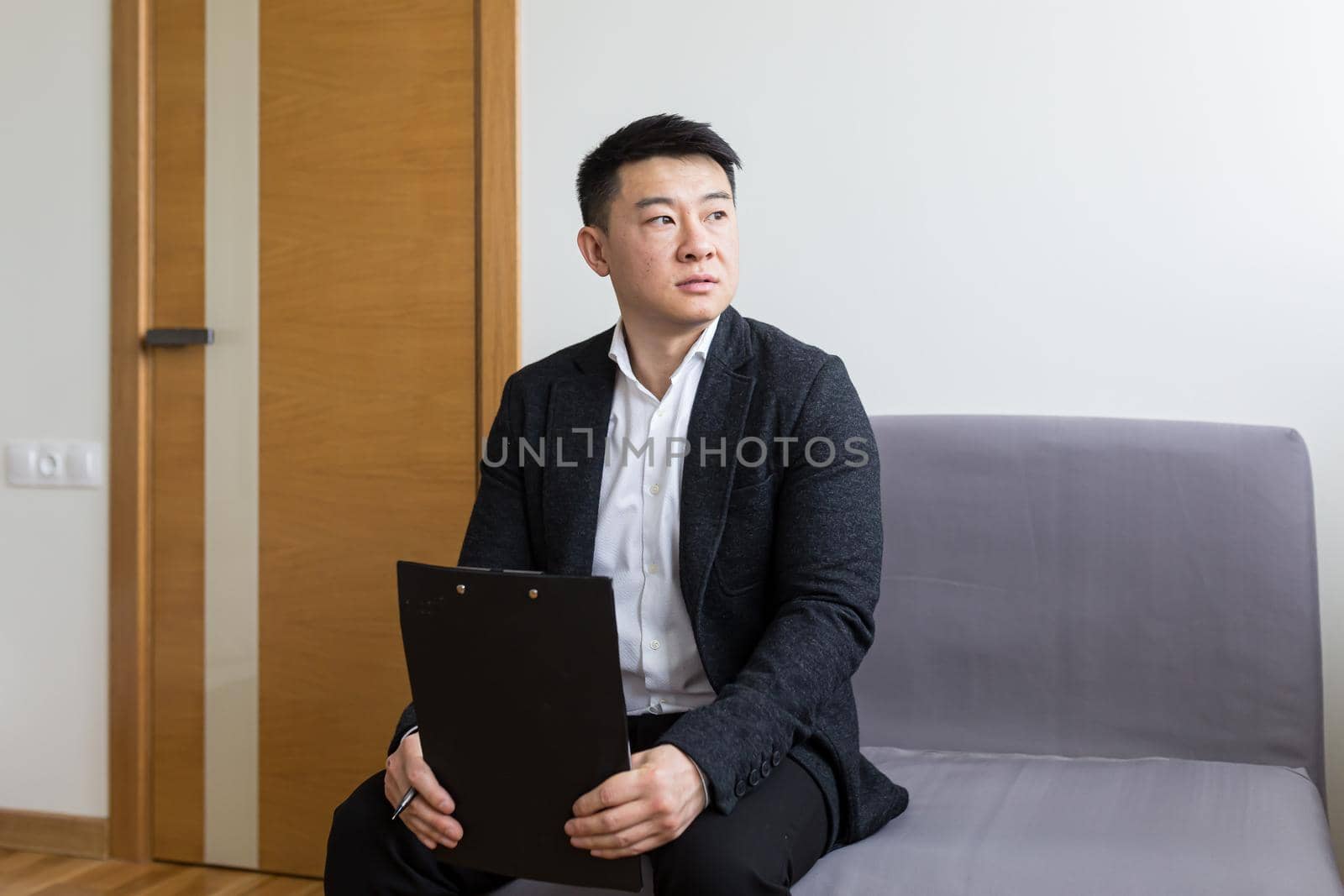 Young successful Asian man, waiting for a job interview, in the waiting room of the office center, sitting on a chair near the reception of the office center