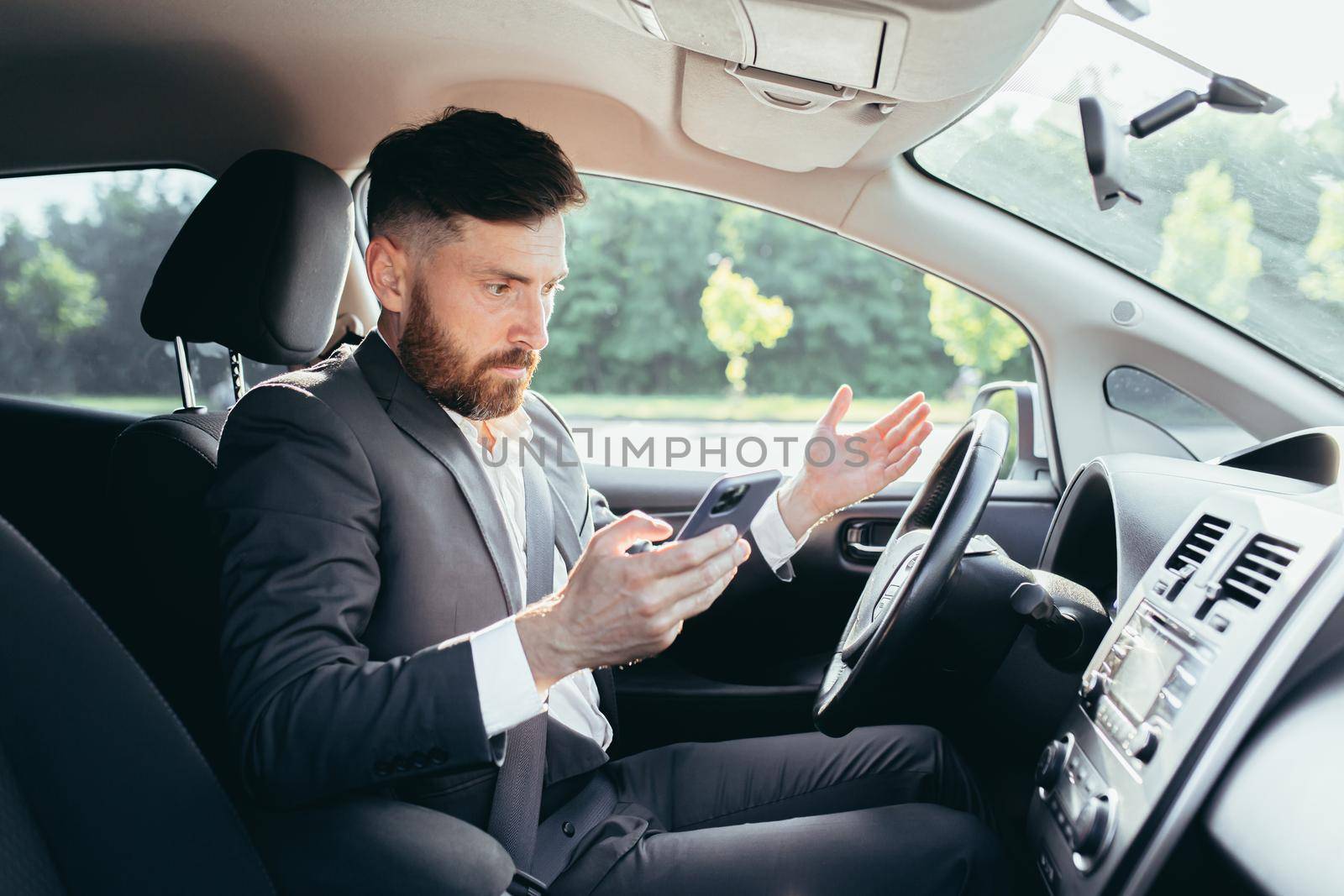 Male businessman sitting behind the wheel of a car, frustrated can not understand the steering and instructions funky car, reads additional information from the phone
