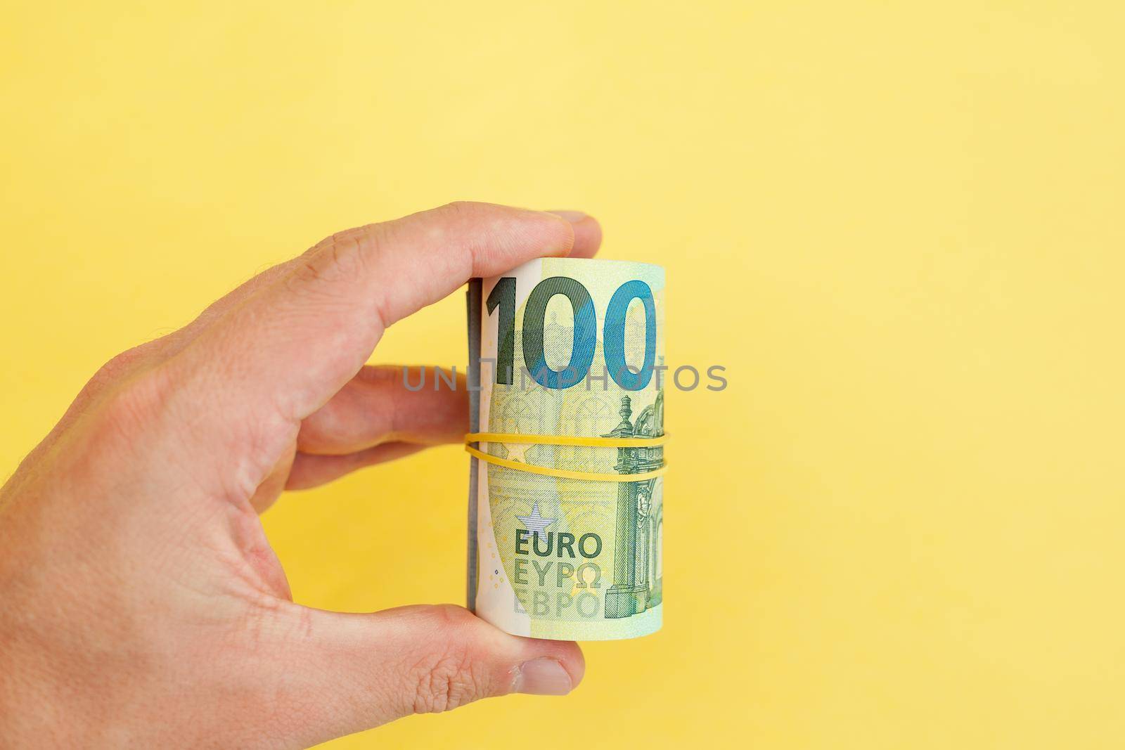 The man holds in his hand money roll of euro banknotes with yellow rubber band on yellow background. cash paper currency, payment, earning and savings, money and finance concept. Empty space for text