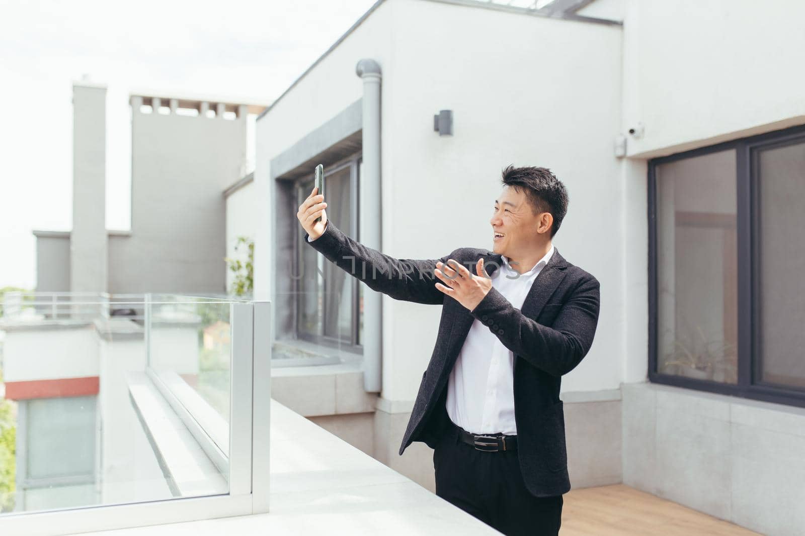 Successful asian businessman man talking on video phone, standing on balcony terrace of modern office center, happy and smiling