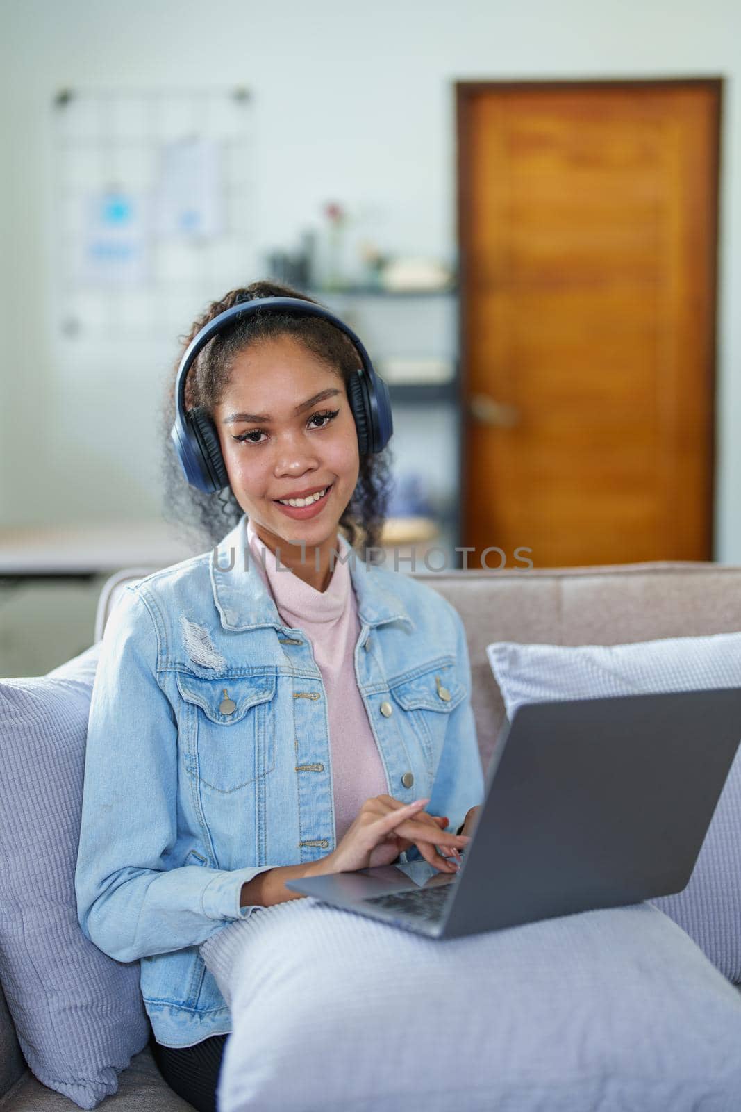 Portrait of an African American sitting on the sofa wearing on-ear headphones and using a computer at home by Manastrong