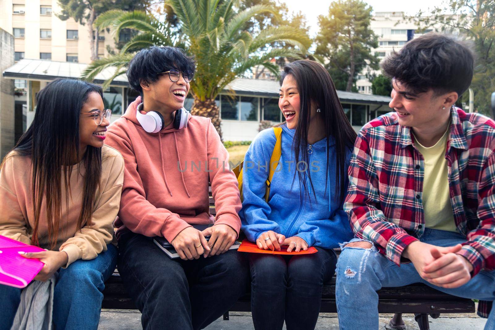 Group of multiracial gen z college student friends talk and laugh sitting on campus bench outside. by Hoverstock