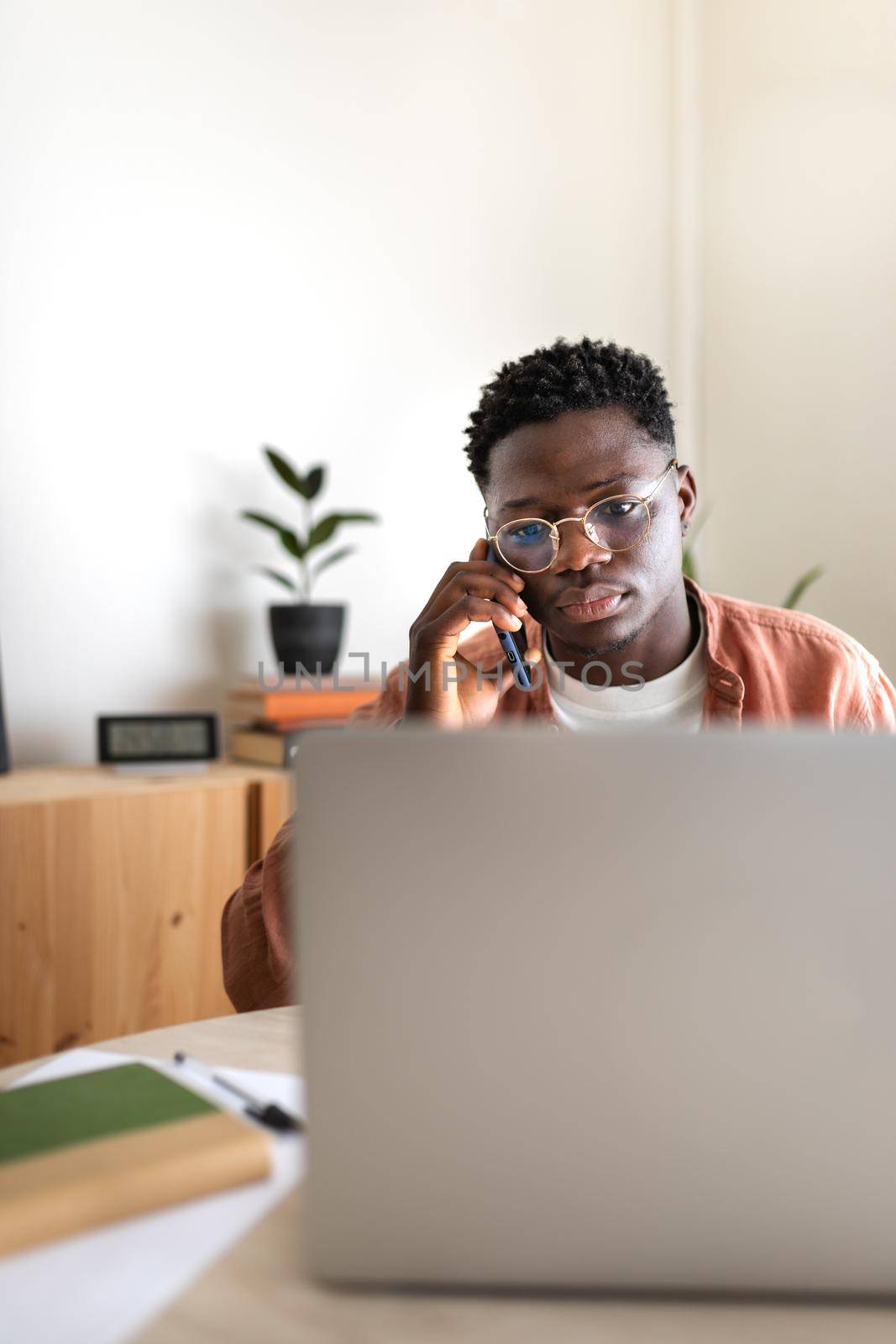 Black male entrepreneur working with laptop at home office talking with mobile phone. Copy space. Vertical. by Hoverstock