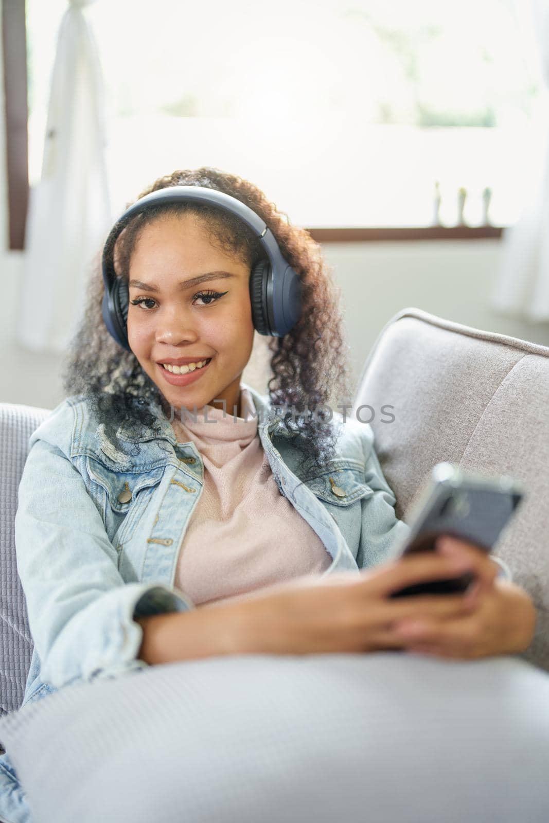 Portrait of an African American sitting on a sofa using a phone and wearing headphones to relax by Manastrong