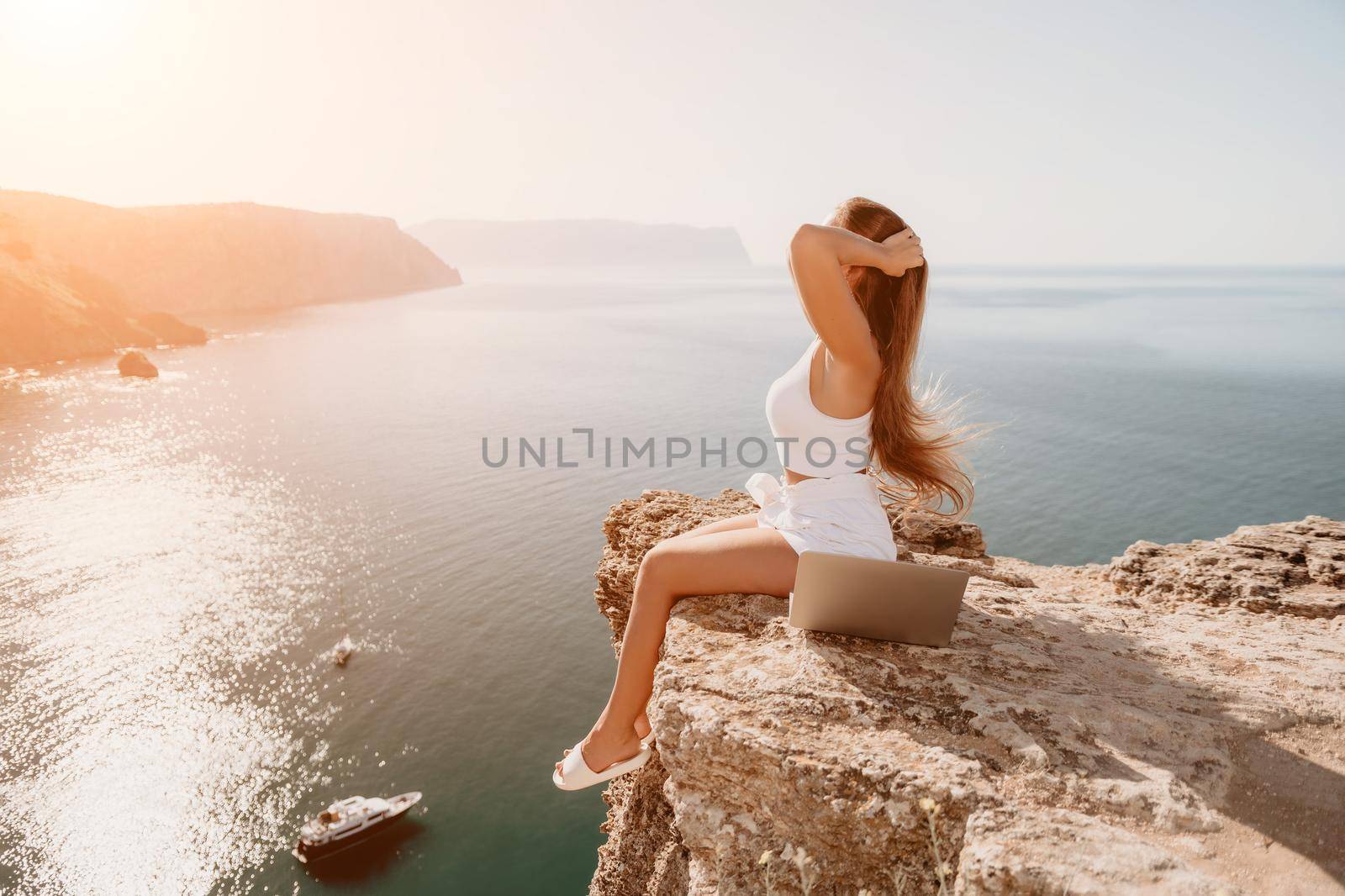 Digital nomad, woman in the hat, a business woman with a laptop sits on the rocks by the sea during sunset, makes a business transaction online from a distance. Freelance, remote work on vacation. by panophotograph