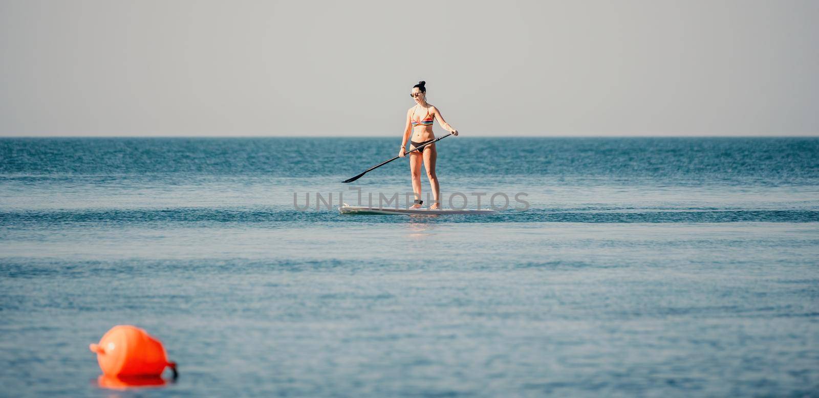 Sea woman sup. Silhouette of happy middle aged woman in rainbow bikini, surfing on SUP board, confident paddling through water surface. Idyllic sunset. Active lifestyle at sea or river. by panophotograph