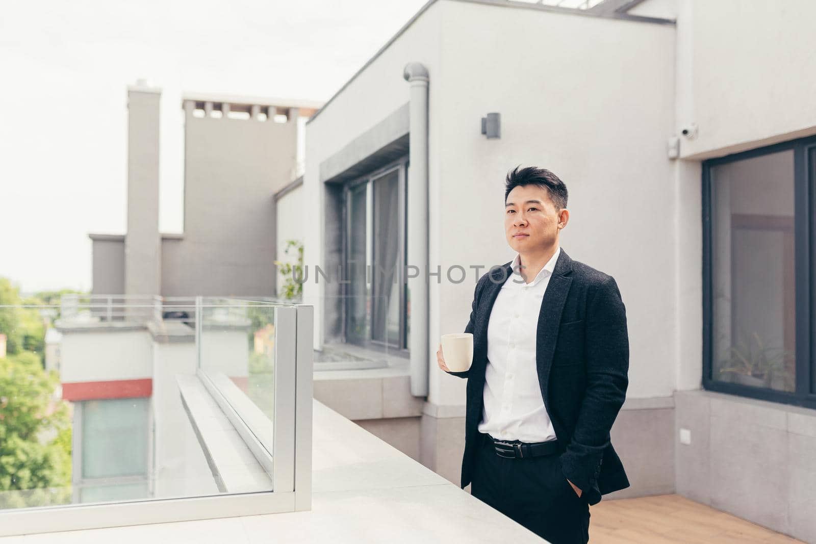 Young successful Asian businessman in a business suit drinking tea or coffee standing on the balcony or terrace of a modern office center