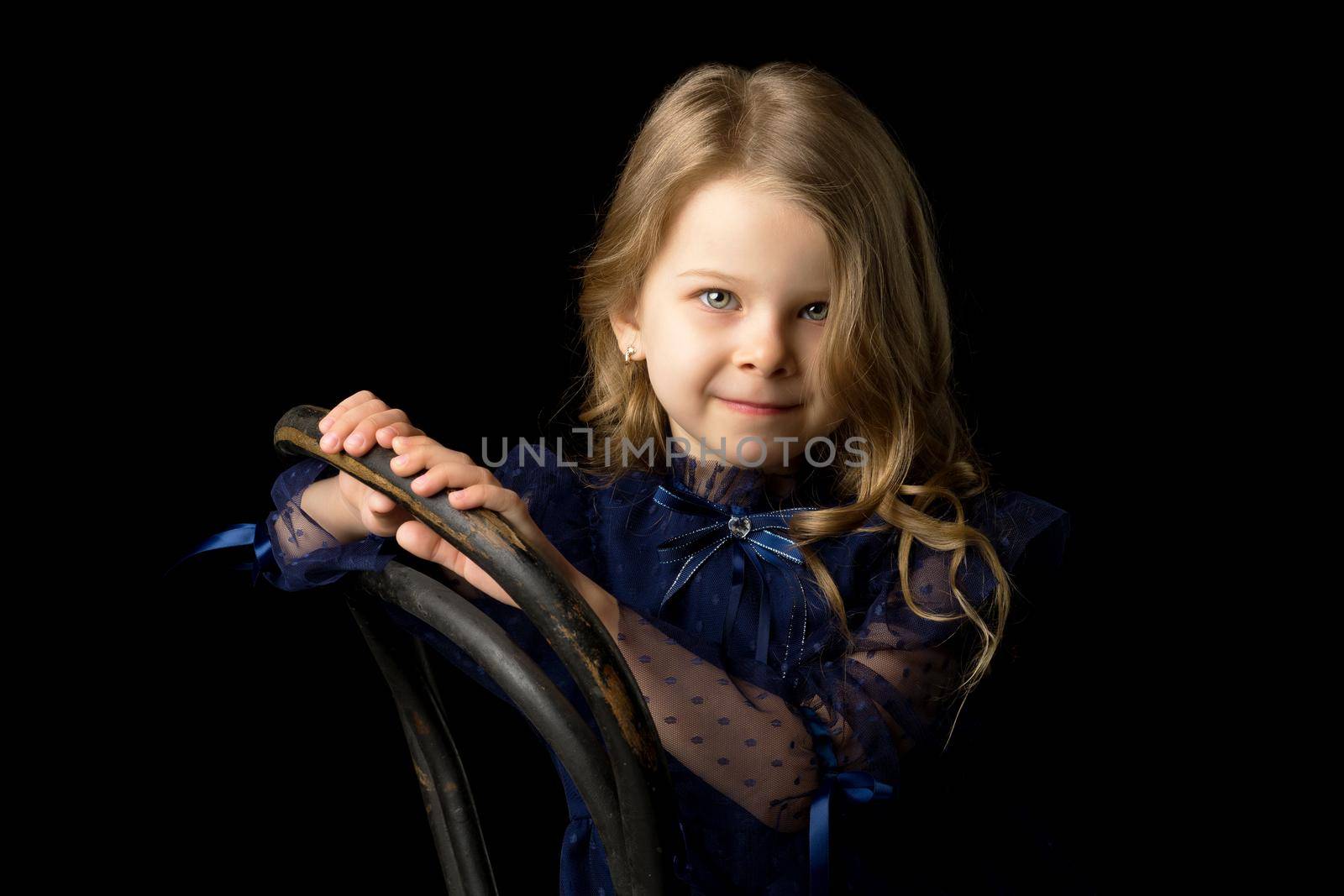 Close up portrait of happy sitting little girl. Lovely smiling blonde girl wearing fashionable clothes sitting on chair on isolated black background in studio.