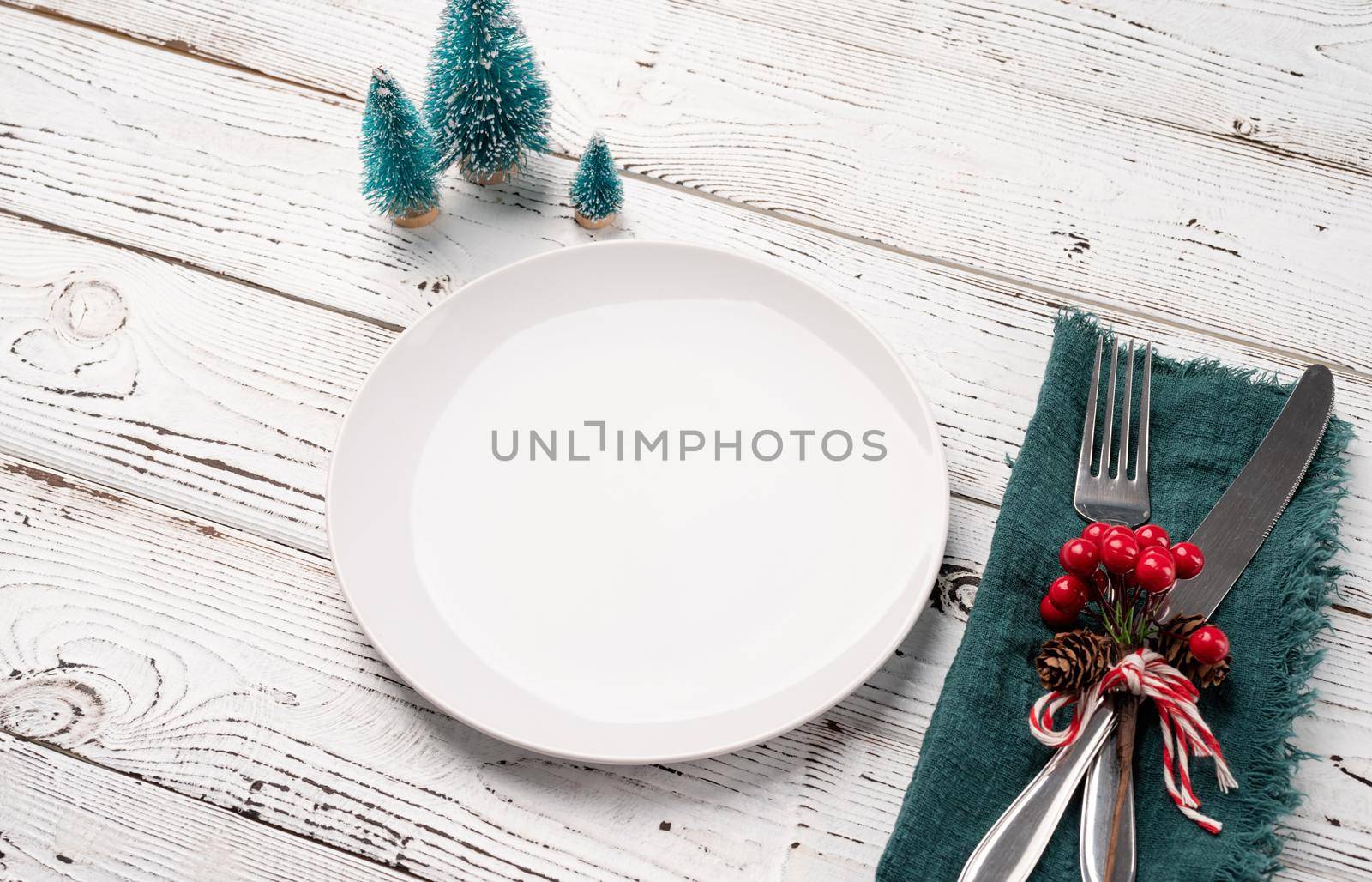 Christmas table setting with white dishware, silverware and red and green decorations on white wooden background high angle view