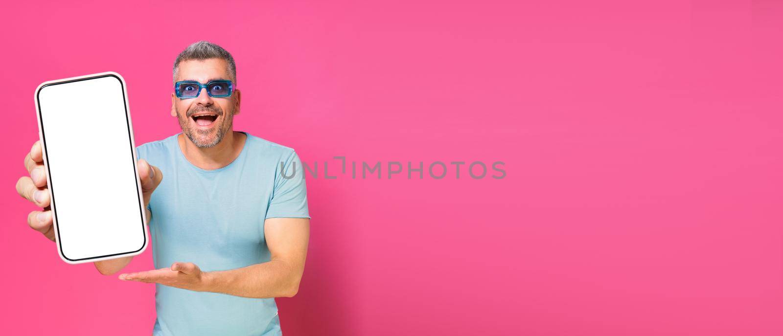 Smartphone in hand of handsome man, guy 30s 40s in casual blue shirt and sunglasses isolated on pink background. Man with phone studio shot. Mobile app advertisement. Copy space.