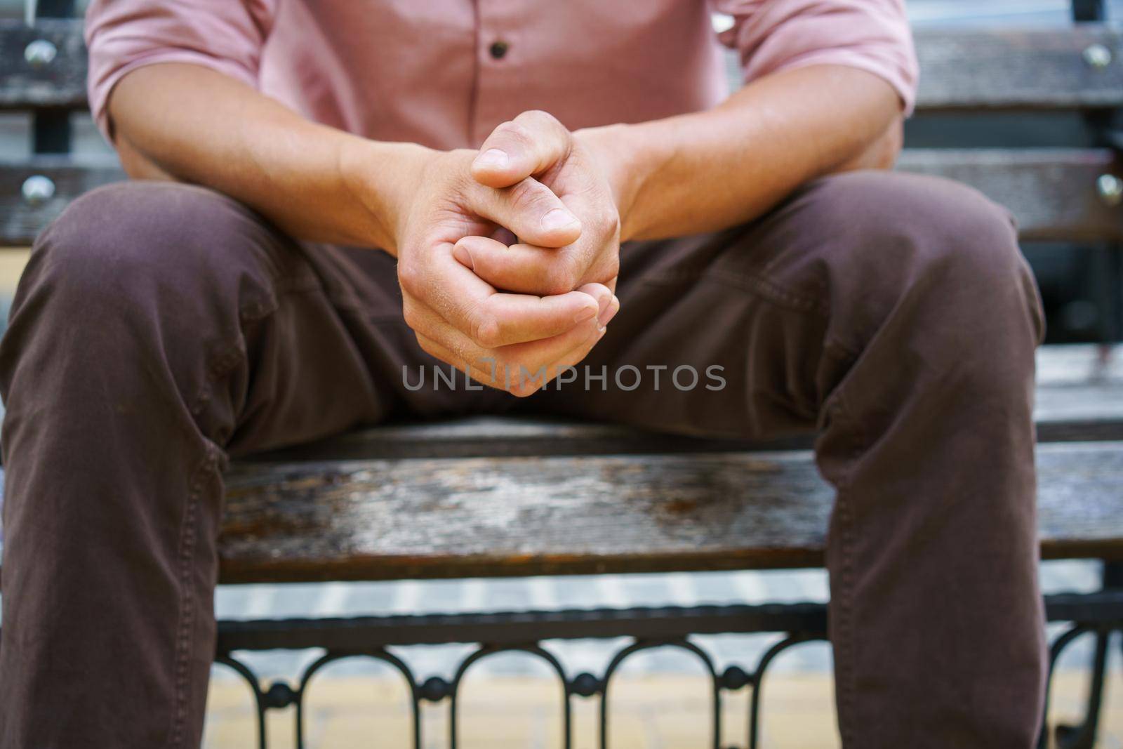 No face visible. Man spend time sitting on the bench with hands put together wearing pink shirt. Overpassing problems young man sits outdoors thinking on life moments. Psychology concept.