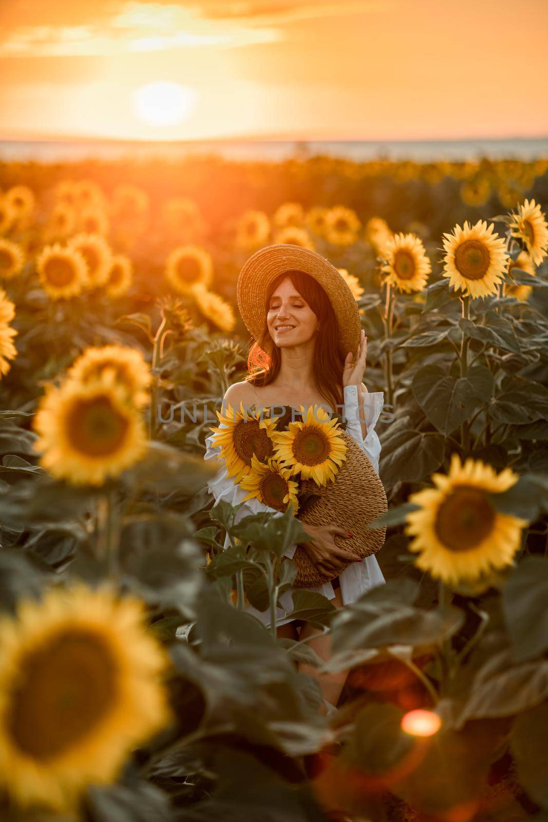 A girl in a hat on a beautiful field of sunflowers against the sky in the evening light of a summer sunset. Sunbeams through the flower field. Natural background