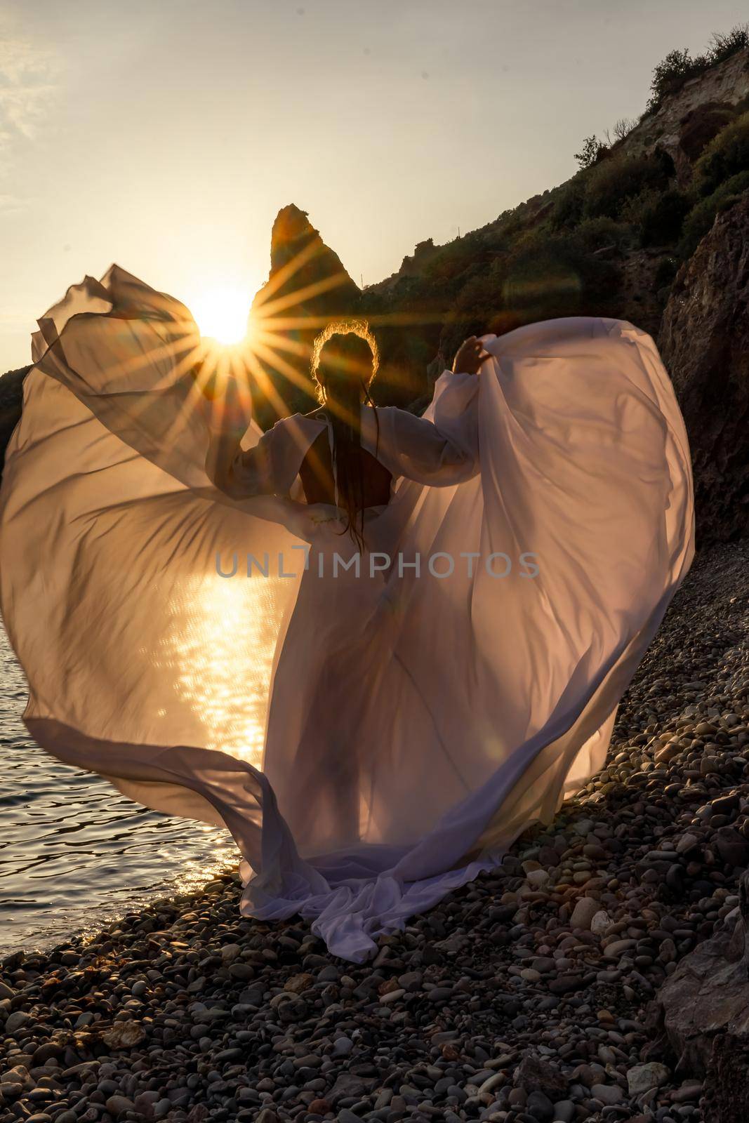 A mysterious female silhouette with long braids stands on the sea beach with mountain views, Sunset rays shine on a woman. Throws up a long white dress, a divine sunset