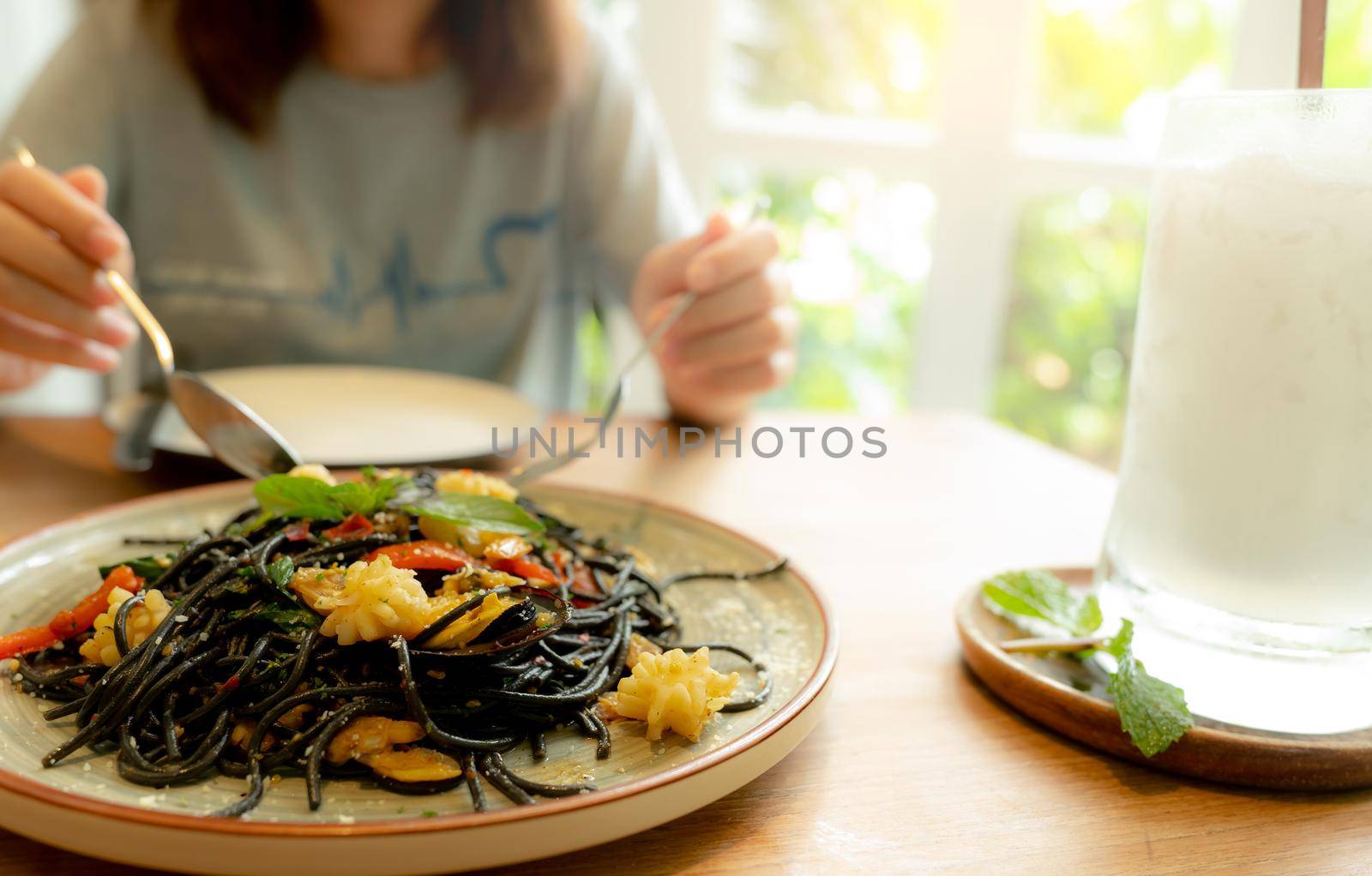 Black cuttlefish ink spaghetti with shrimp on plate. Black pasta with squid ink on a restaurant table and blur woman eating with fork and spoon. Healthy food. Woman eating delicious black spaghetti. by Fahroni