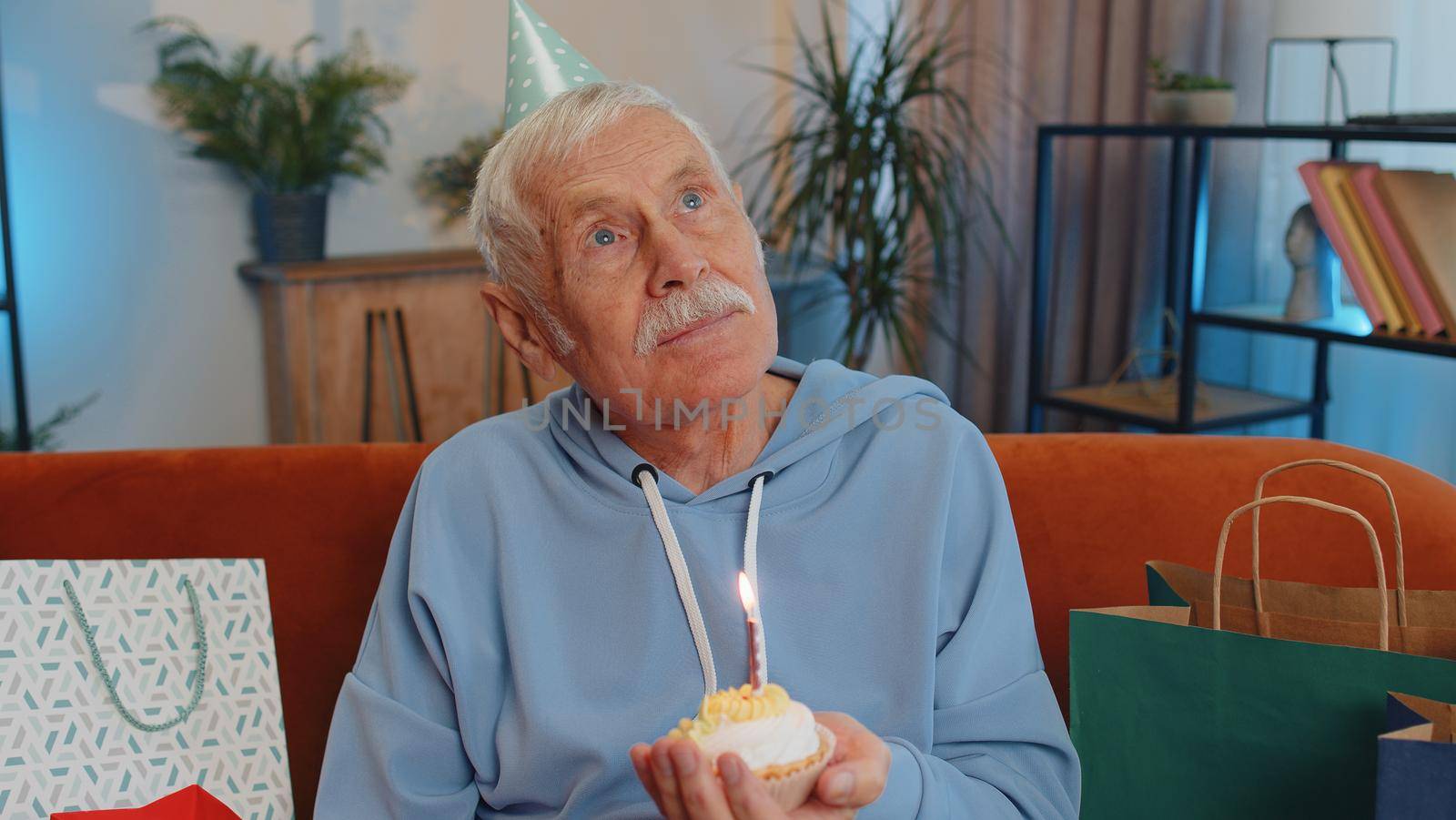 Happy senior grandfather wears festive birthday hat hold cupcake makes wish joyful congratulating blowing burning candle on cake. Mature man celebrating anniversary party on couch with gifts at home