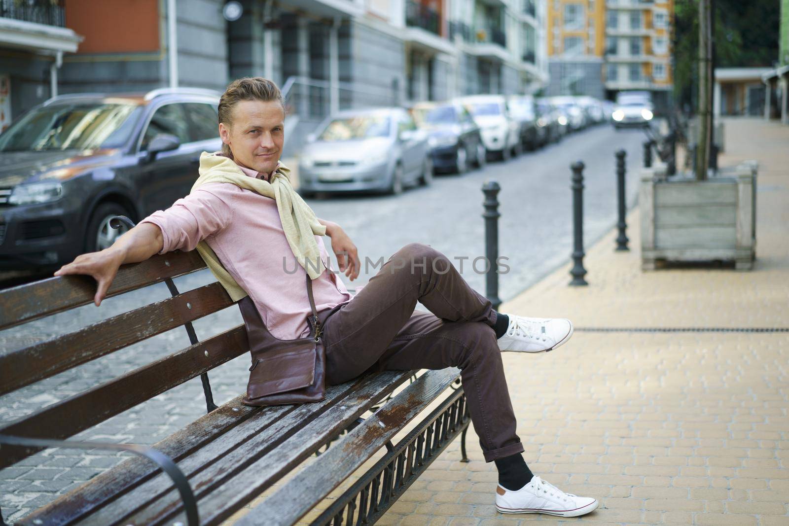 Handsome man sits on the bench with legs crossed spend time in urban city near his office or home, wearing pink shirt and shoulder bag. Freelancer man off work.