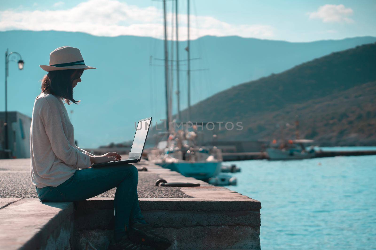 A young girl in a hat is sitting on the pier and working, typing on a laptop keyboard on a sunny day against a beautiful background of a seascape with moored yachts in the bay. Woman works and travels