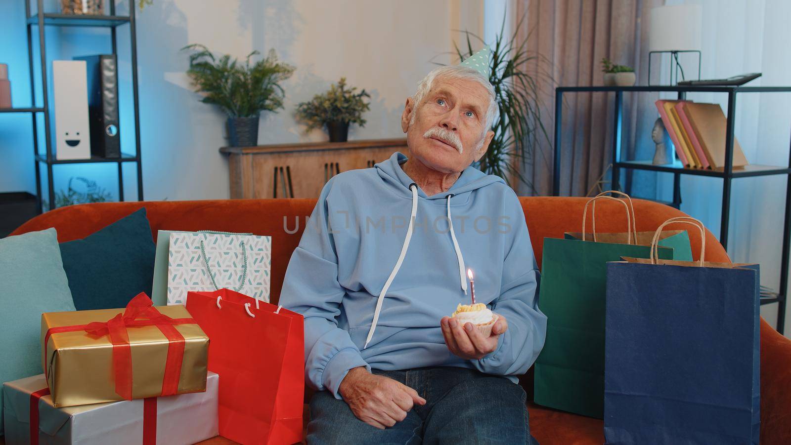 Happy senior grandfather wears festive birthday hat hold cupcake makes wish joyful congratulating blowing burning candle on cake. Elderly man celebrating anniversary party on couch with gifts at home