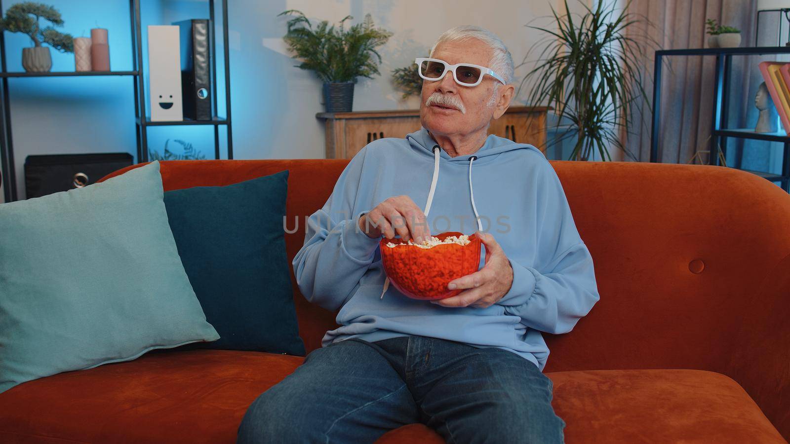 Excited elderly man on sofa eating popcorn and watching interesting tv serial, sport game, film, online social media movie content online at home. Senior grandfather enjoying domestic entertainment
