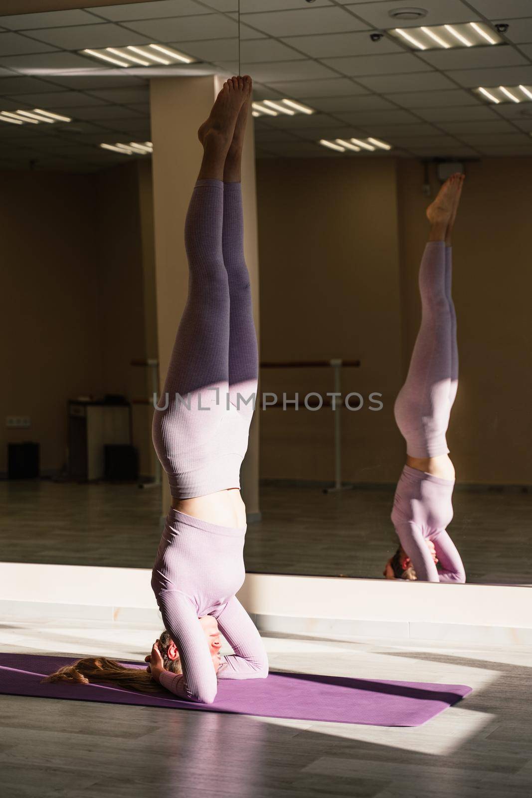 A slender peaceful positive Caucasian fitness model in a purple suit stands on her head on a purple rug. A woman performs S hirshasana. The concept of body positivity, healthy lifestyle.