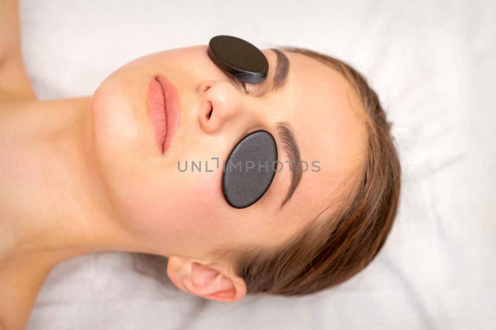 Young woman receiving facial massage with black mineral stones on her eyes in a spa