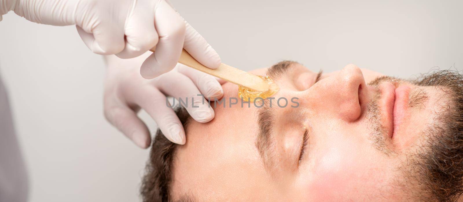 Beautician applying wax paste between eyebrows during the procedure of waxing in the beauty salon