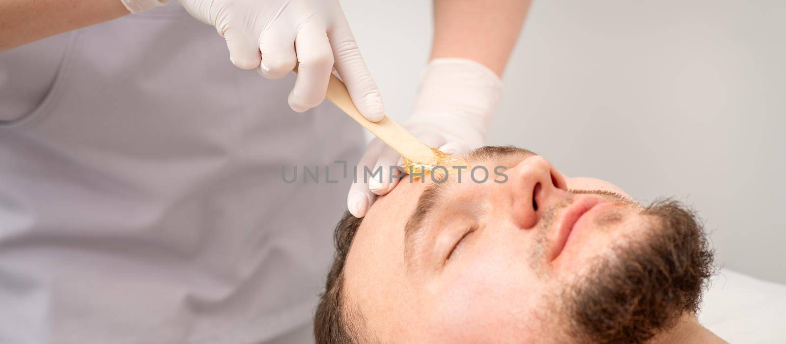 Beautician applying wax paste between eyebrows during the procedure of waxing in the beauty salon. by okskukuruza