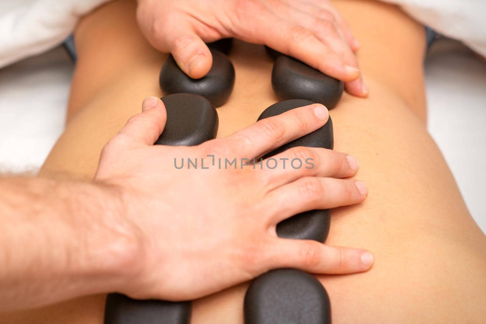Young caucasian woman receiving back massage with black stones by masseur in spa salon. A woman getting a spa treatment. by okskukuruza