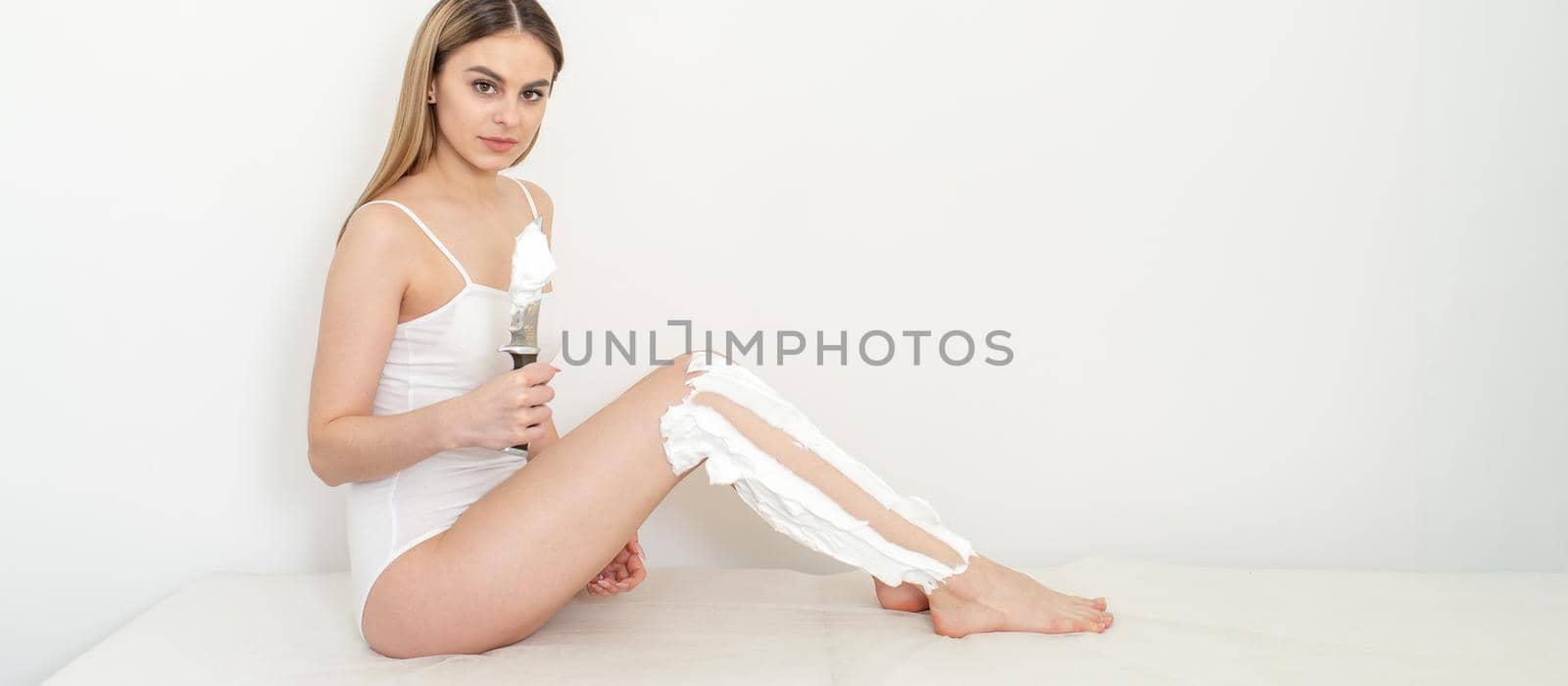 Beautiful caucasian woman shaving her legs with a knife with shaving foam on white background. Depilation and epilation concept. by okskukuruza