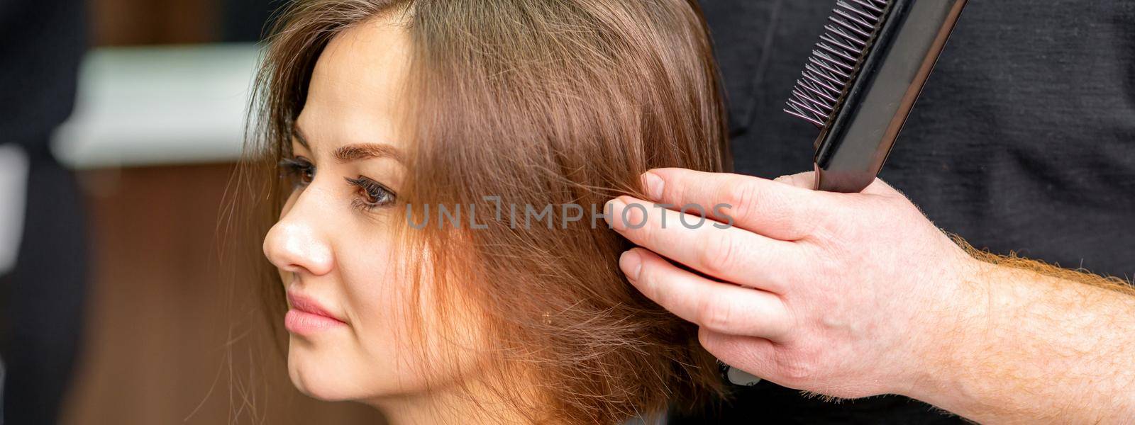 Male hairdresser works on the hairstyle of the young caucasian brunette woman at a hair salon