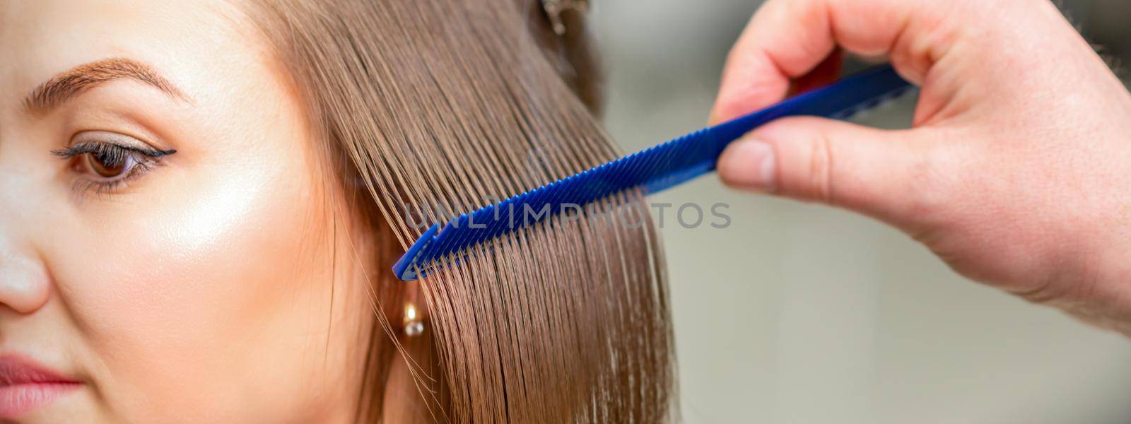 Hairdresser straightens female brown hair of medium length with a hair with an iron hair straightener and comb in a beauty salon. by okskukuruza