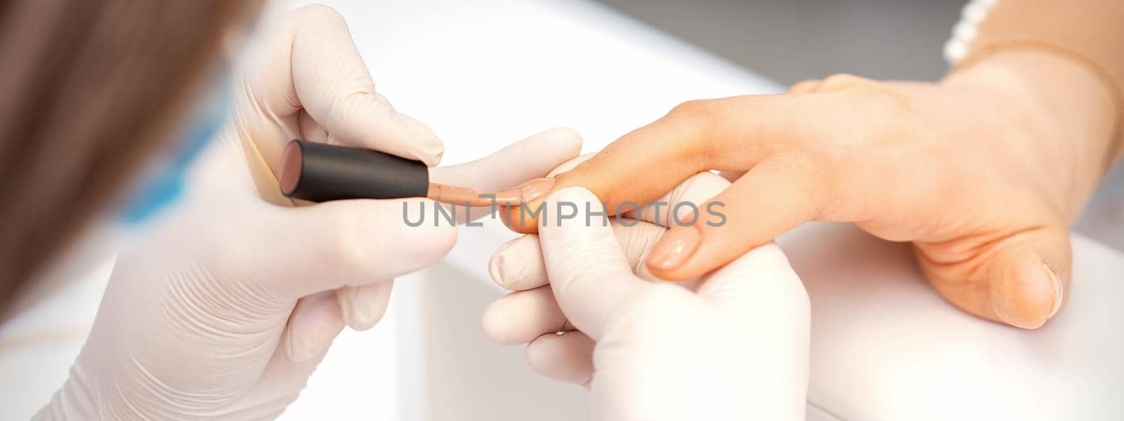 Hands of manicure master in white protective gloves apply polish to female fingernails. by okskukuruza