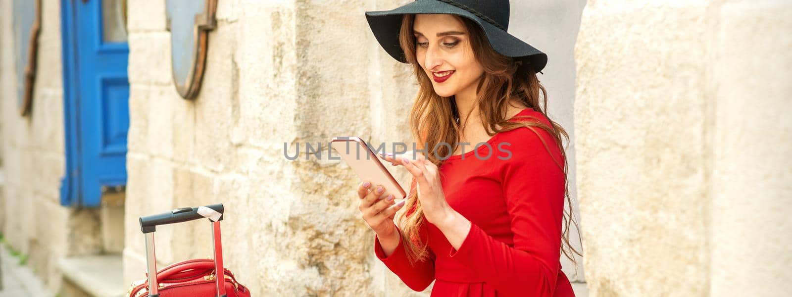 A young traveling white woman in the red jacket and black hat sitting with a smartphone on the city street