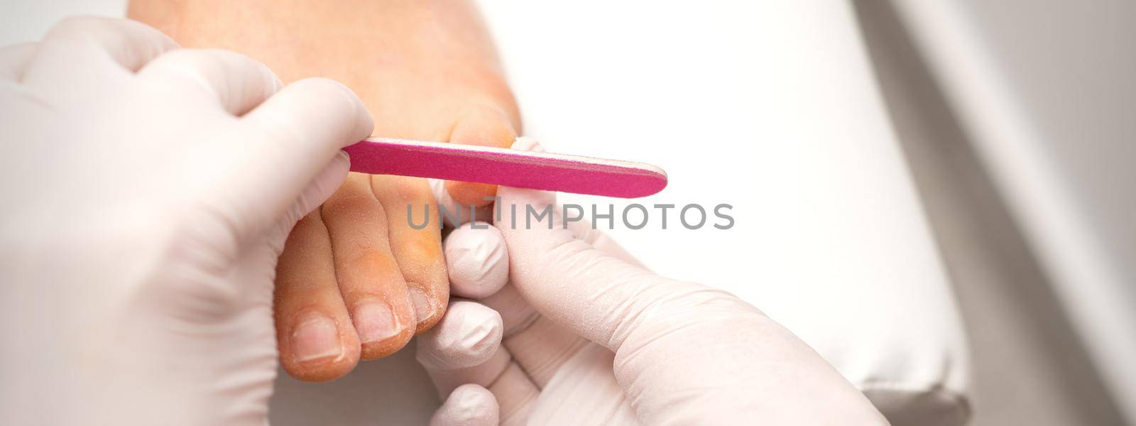 Pedicurist hands in protective rubber gloves filing toenails on feet with a nail file in a beauty salon