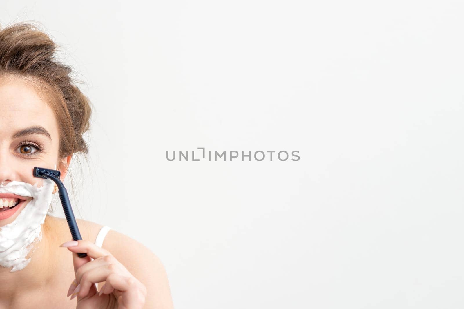 Beautiful young caucasian woman shaving her face by razor on white background. Pretty smiling woman with shaving foam and razor on her face