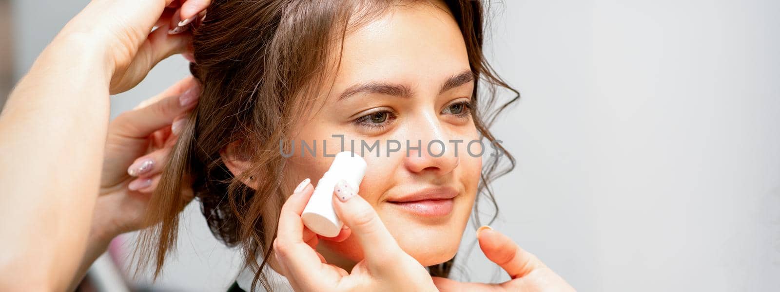 Makeup artist applying cream blush foundation tube on the cheek of the young caucasian woman in a beauty salon