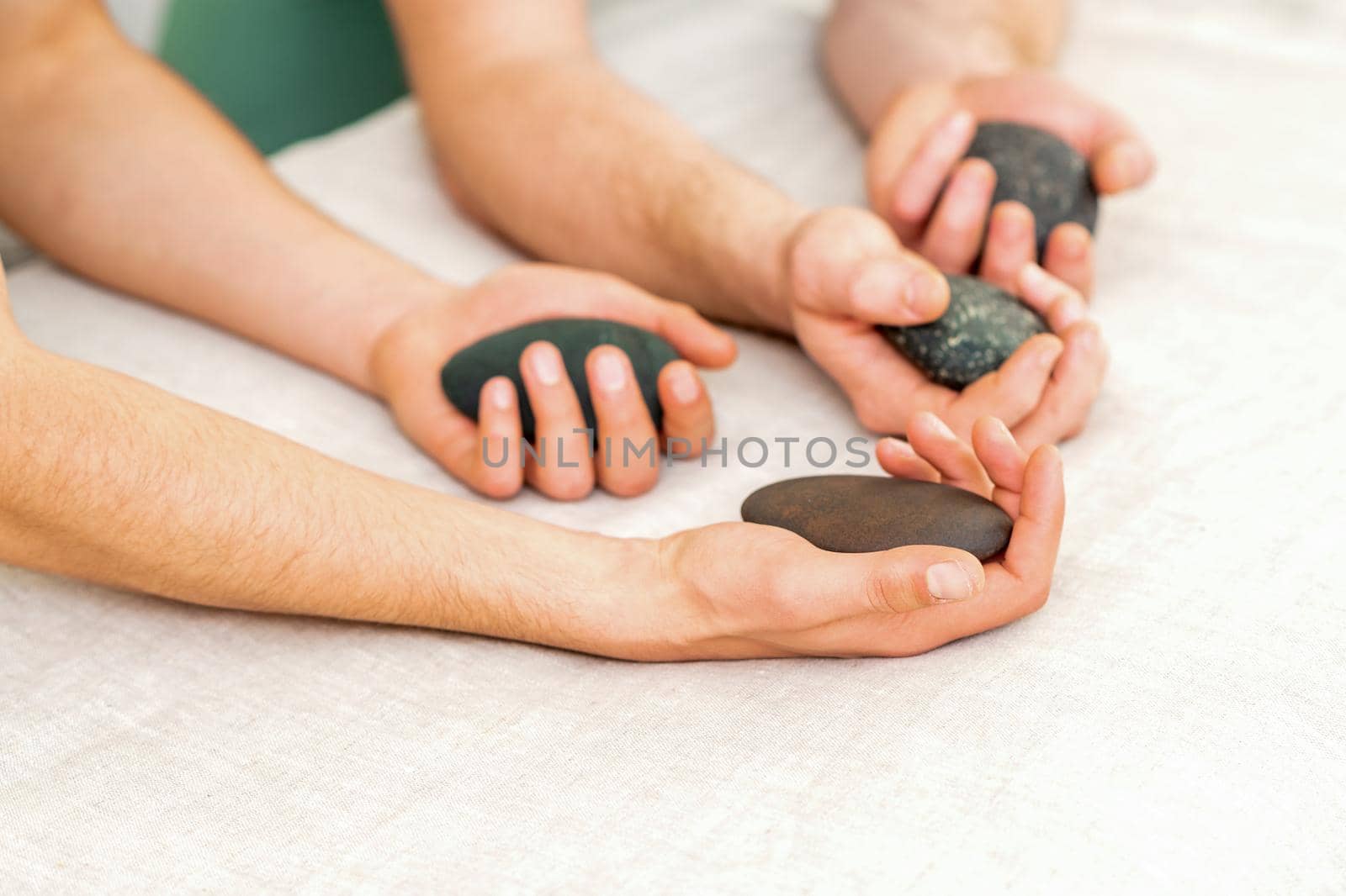 Four hands of male masseurs holding spa stones on white towel top view. Stone massage concept