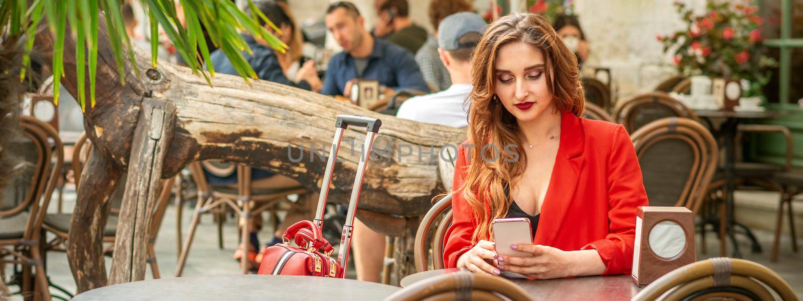 Tourist young caucasian white woman in a red jacket looking at smartphone sitting at the table in cafe outdoors