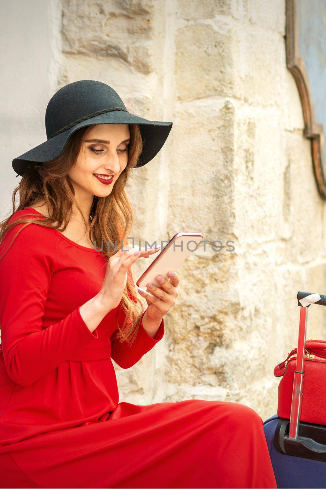 A young traveling white woman in the red jacket and black hat sitting with a smartphone on the city street