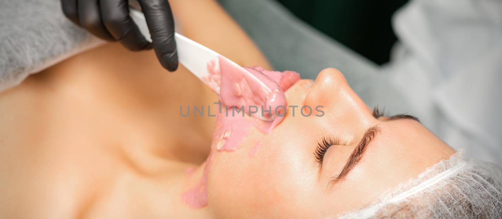 Beautician applying alginate peel-off powder facial mask with the spatula in a spa. by okskukuruza