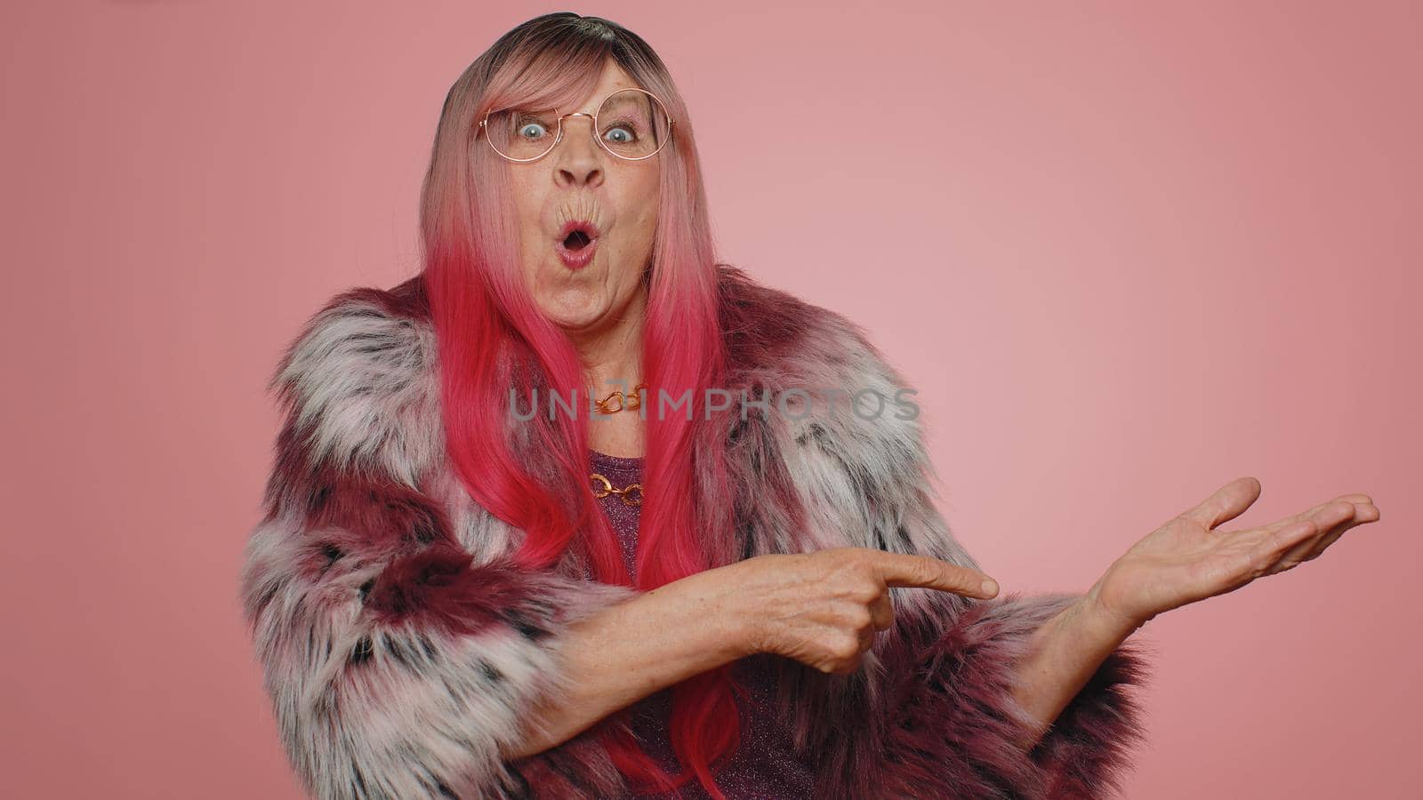 Lovely old woman showing thumbs up and pointing empty place, advertising area for commercial text, copy space for goods promotion. Senior elderly grandmother. Indoors studio shot on pink background
