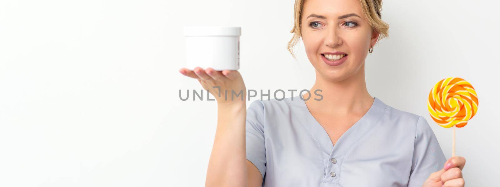 Beautician holding a jar of wax for waxing and candy on a stick smiling on a white background. Natural product for hair removal