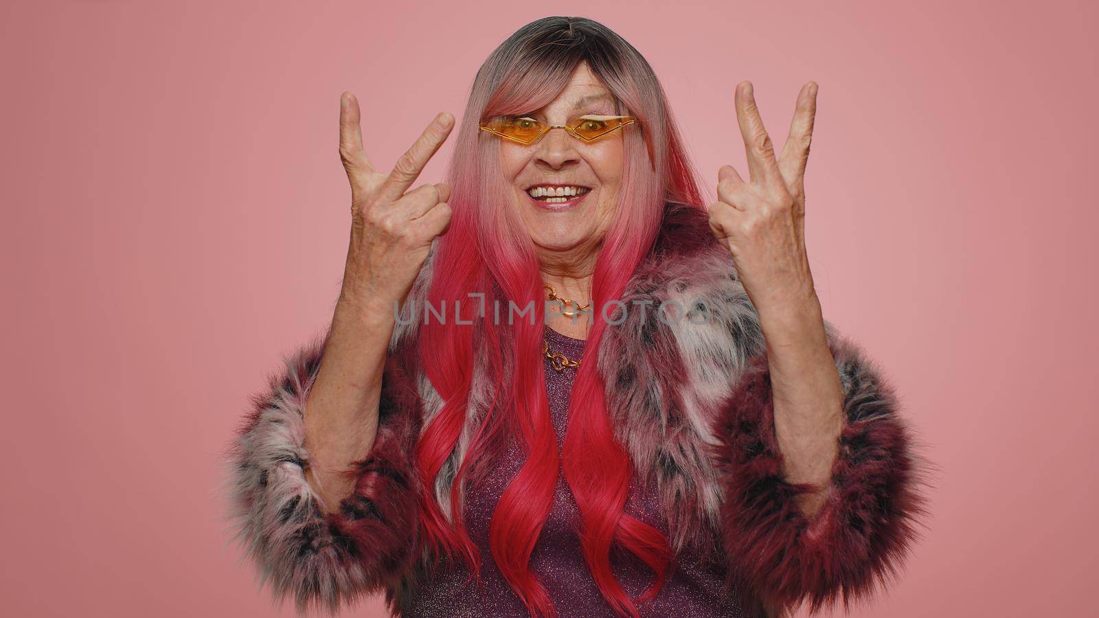 Happy joyful old woman in coat shouting, raising fists in gesture I did it, celebrating success, winning, birthday, lottery goal achievemen. Elderly grandmother indoors on pink studio wall background