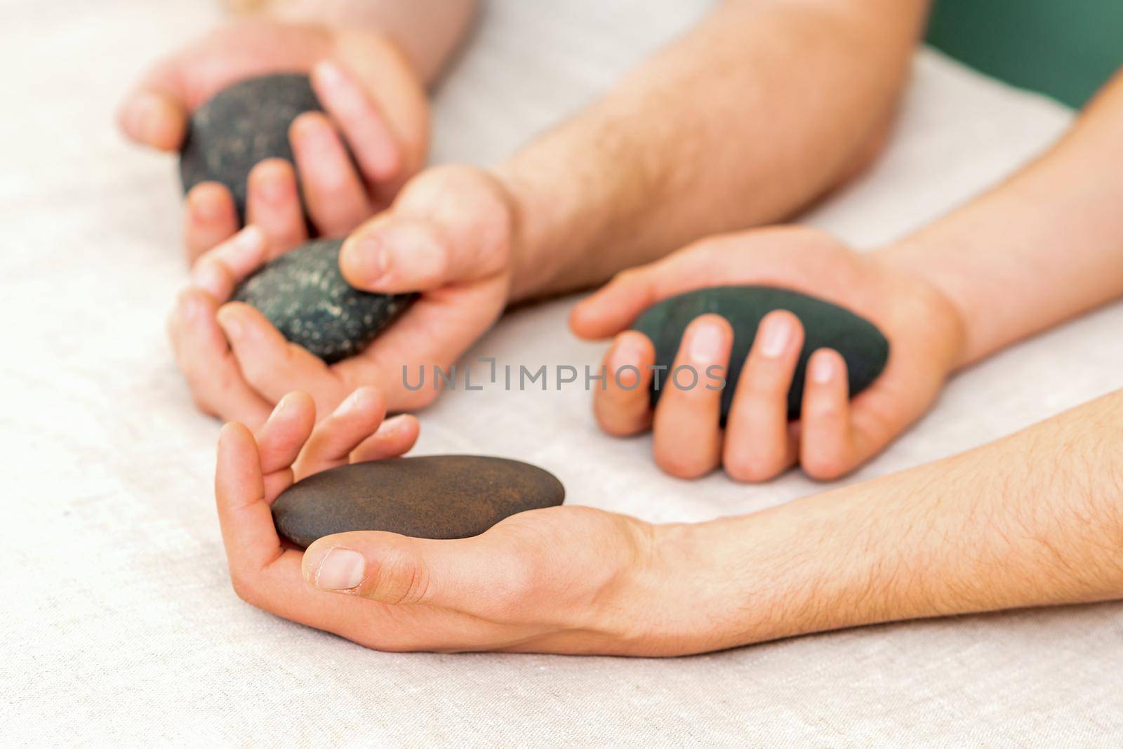 Four hands of male masseurs holding spa stones on white towel top view. Stone massage concept