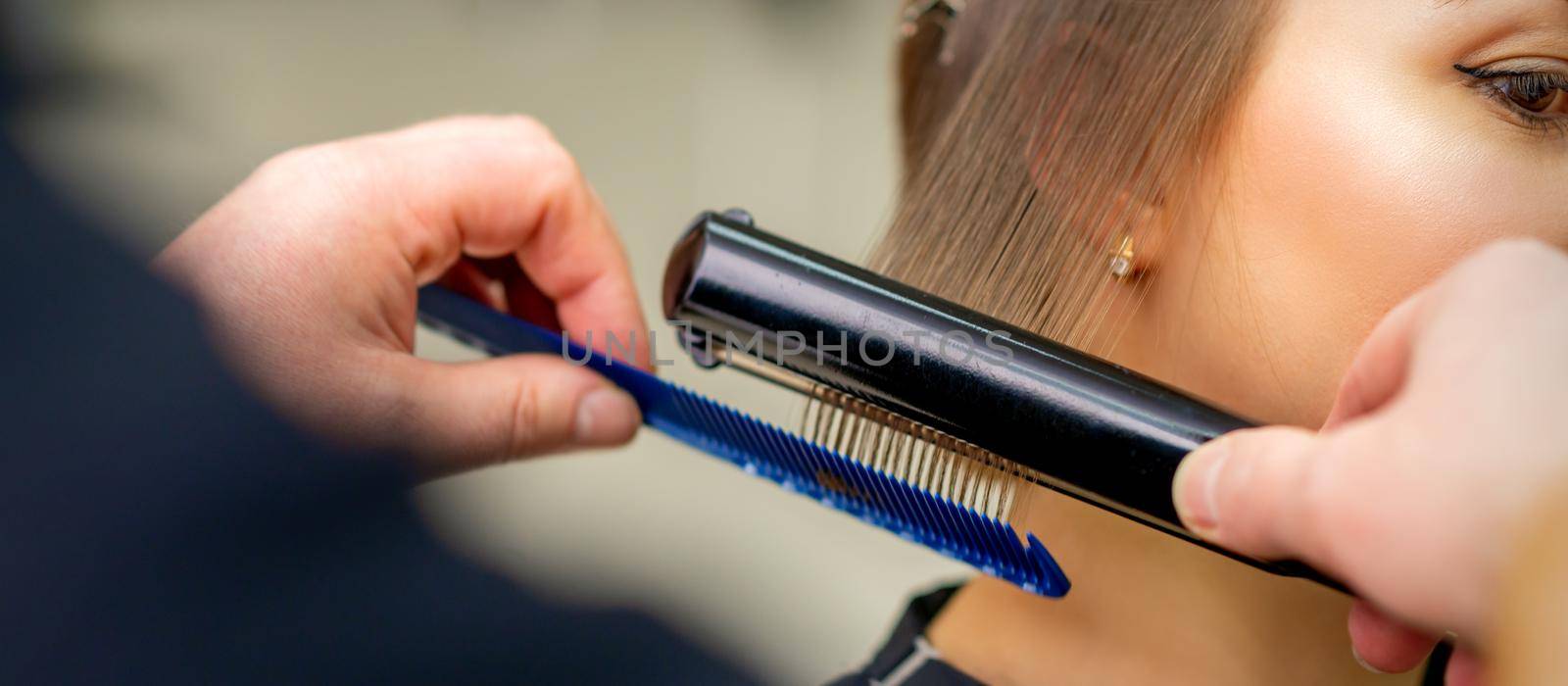 A hairdresser is straightening the hair of the young brunette woman in a beauty salon