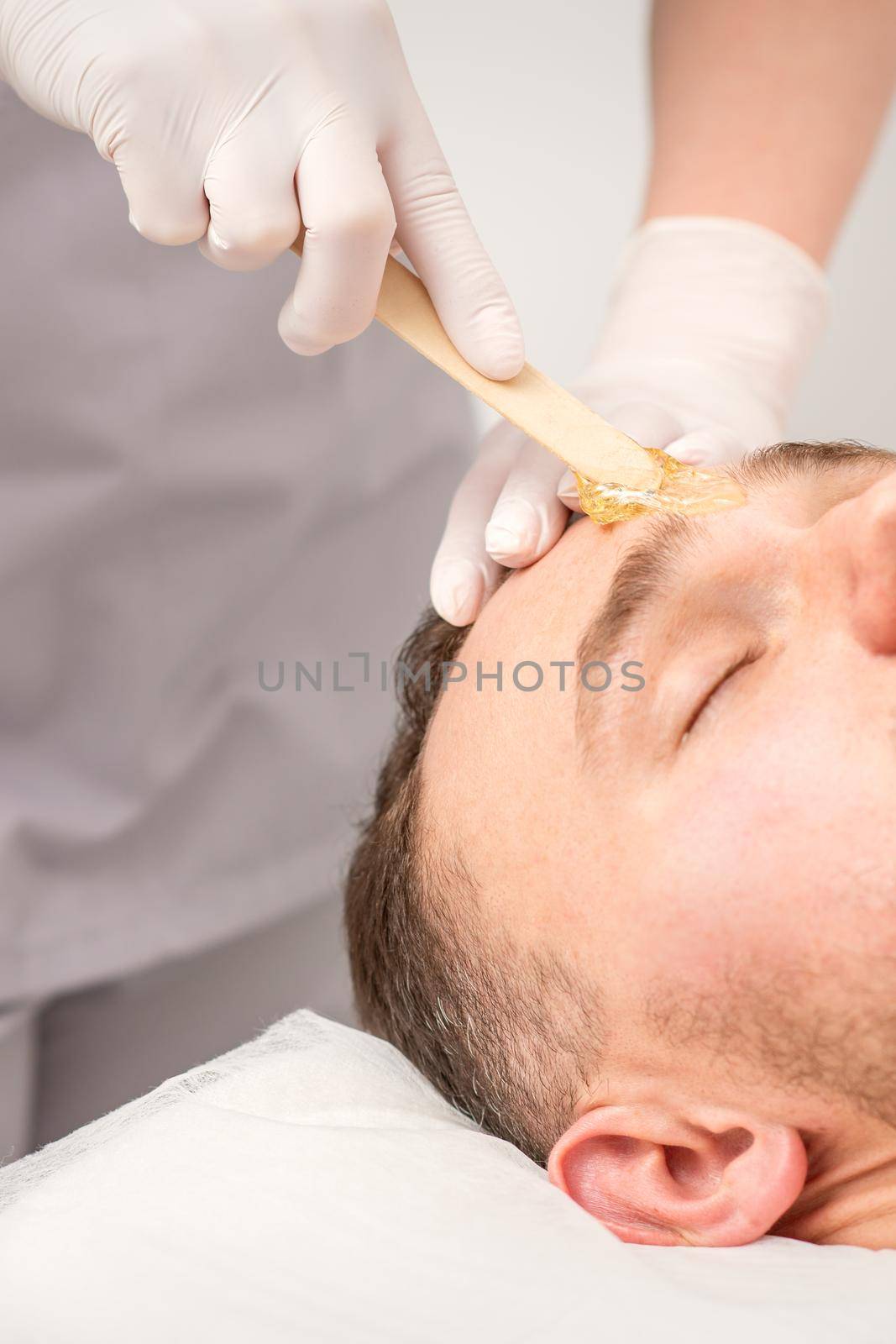 Beautician applying wax paste between eyebrows during the procedure of waxing in the beauty salon