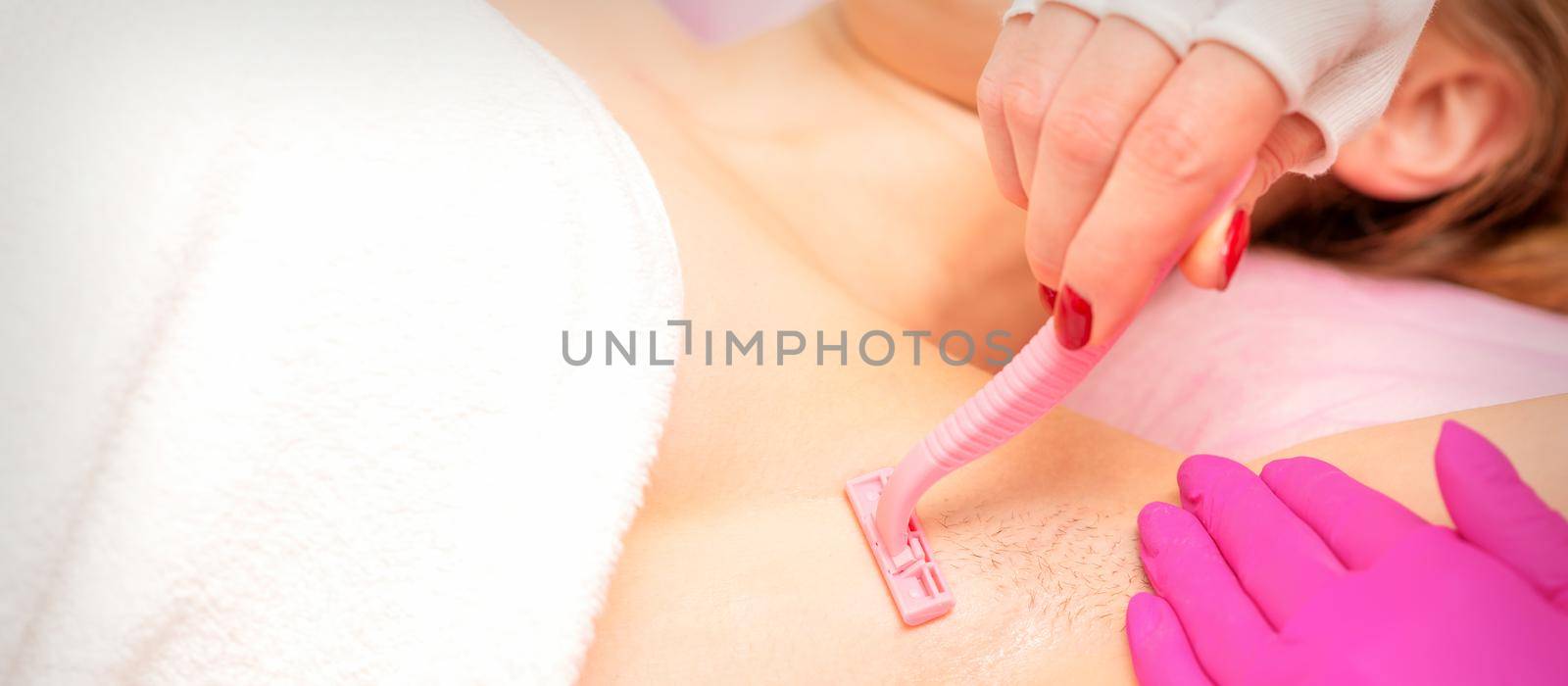 Beautician shaves the armpit of a young woman with a razor before the hair removal procedure